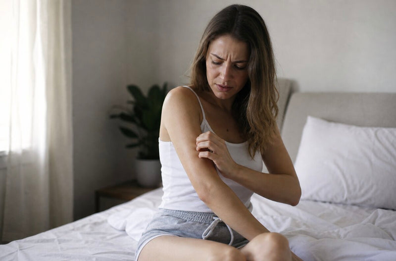 A woman sitting on the edge of her bed, gently scratching her arm with a confused expression, no visible rash